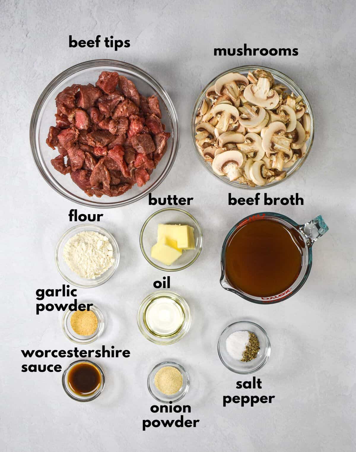 The ingredients for the dish prepped and arranged in glass bowls on a white table with each labeled with small, black letters.