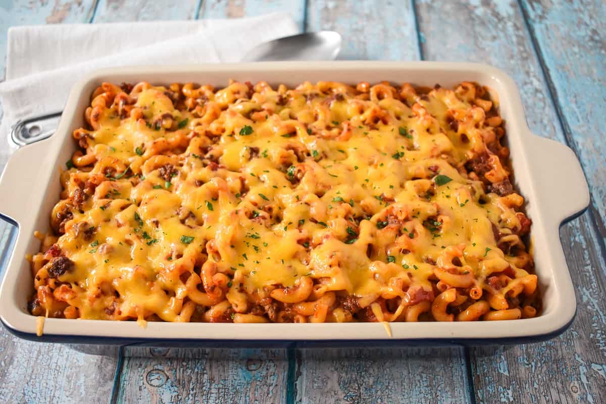 The macaroni and beef casserole in a white baking dish with a serving spoon and white linen in the background.