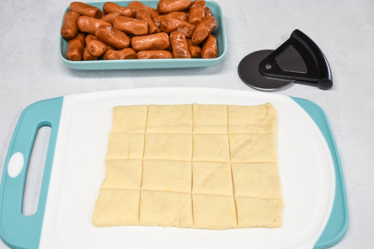 Dough on a cutting board cut into sixteen pieces with a blue platter of cocktail sausage and a small pizza cutter in the background.