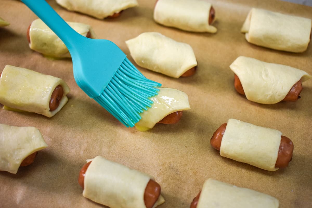 A blue pastry brush applying egg wash to a dough wrapped sausage on a baking sheet.