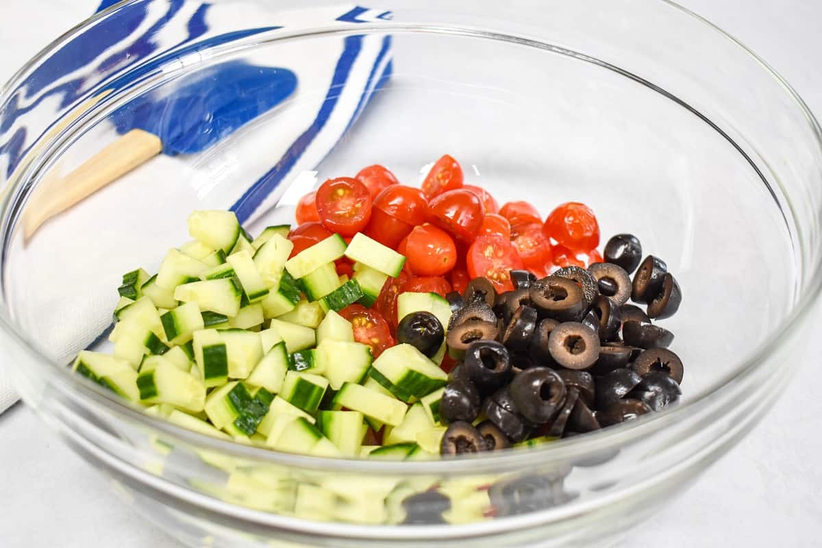 Prepped tomatoes, cucumbers, and black olives in a large, glass bowl.