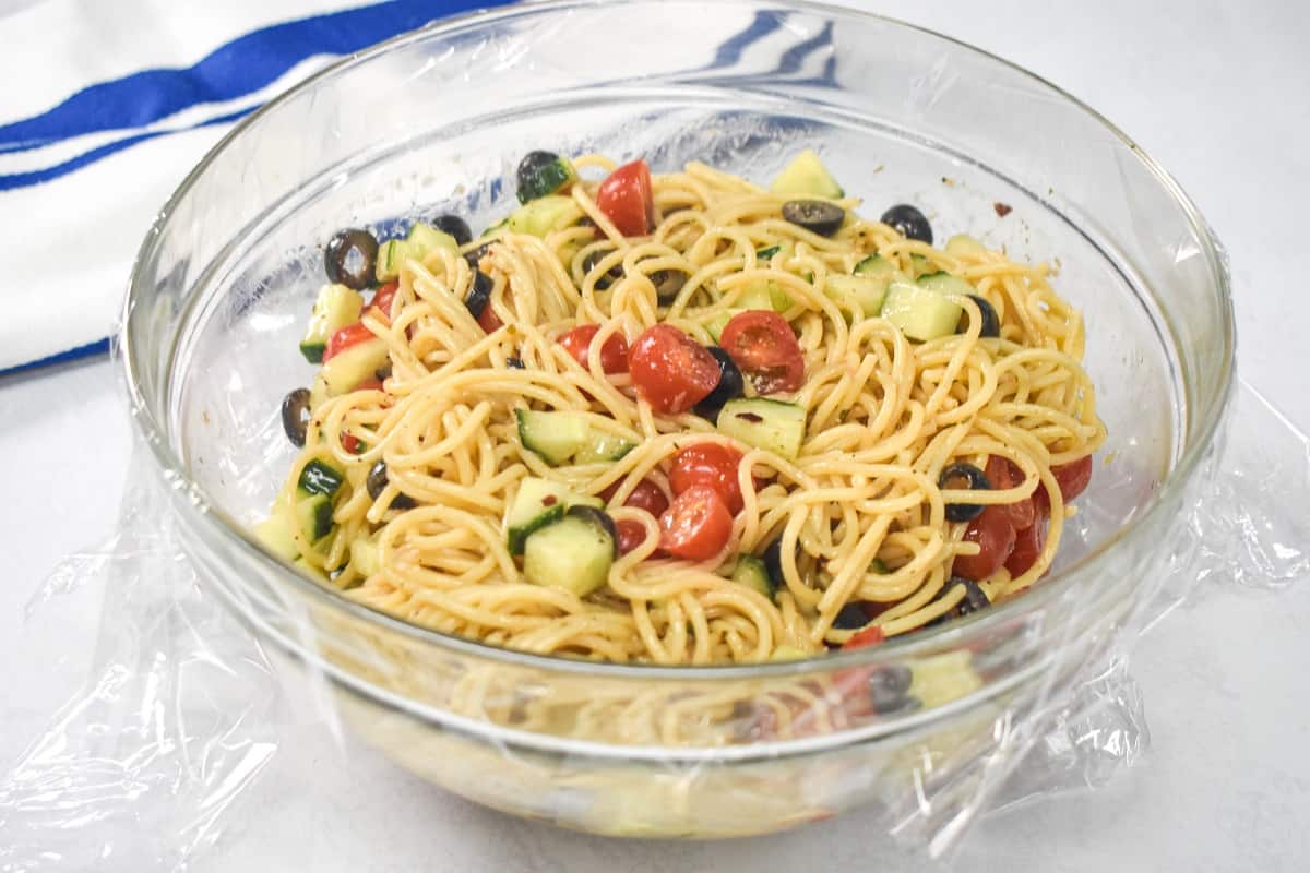 The spaghetti salad in a large glass bowl covered with plastic wrap.