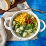 The potato broccoli soup served in a white bowl set on a beige linen on a blue table with sliced bread and a spoon in the background.