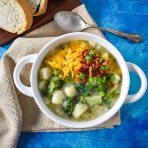 The potato broccoli soup served in a white bowl set on a beige linen on a blue table with sliced bread and a spoon in the background.