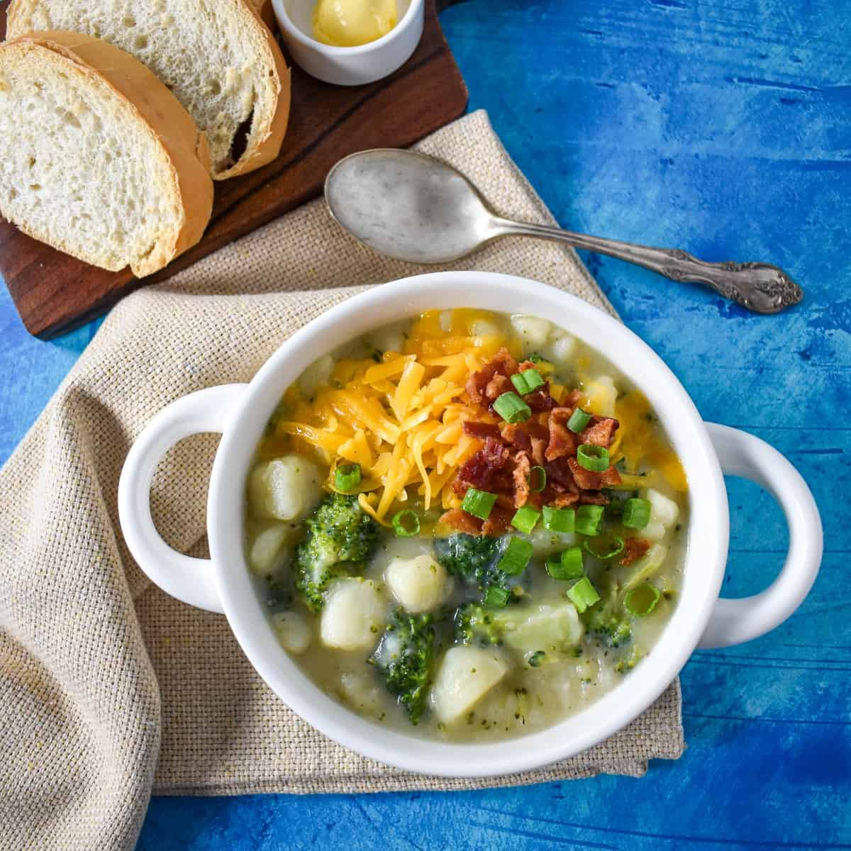 The finished soup served in a white bowl that is set on a beige linen on a blue table with sliced bread and butter on a cutting board and a spoon in the background.