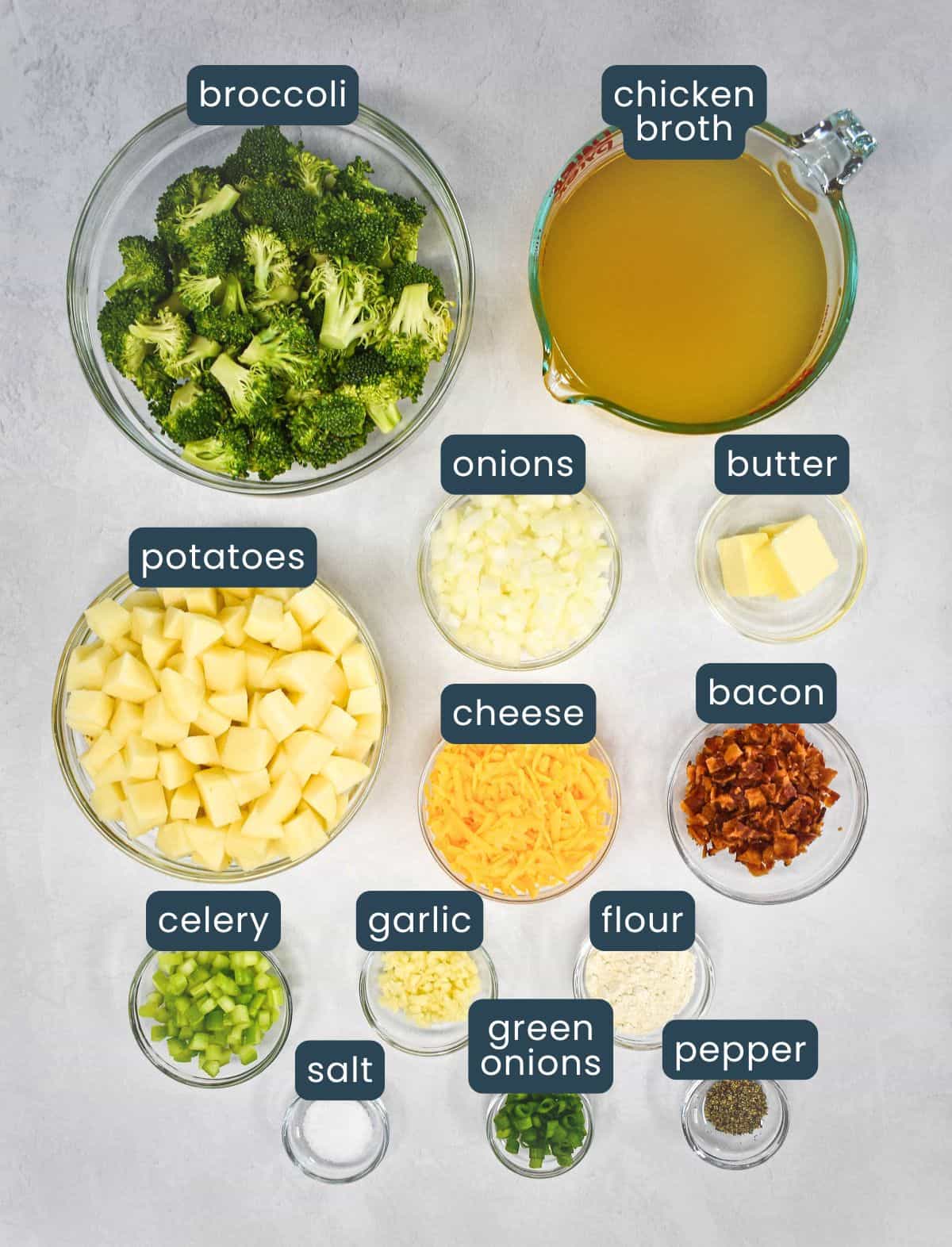 The ingredients for the potato broccoli soup, prepped and arranged in glass bowls on a white table with each one labeled.
