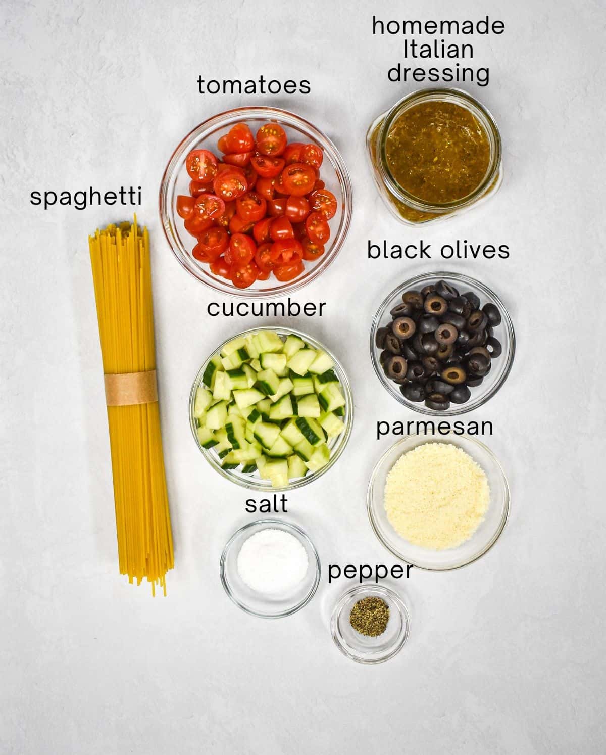 The ingredients for the spaghetti pasta salad prepped and arranged in glass bowls on a white table with each labeled with small, black letters.