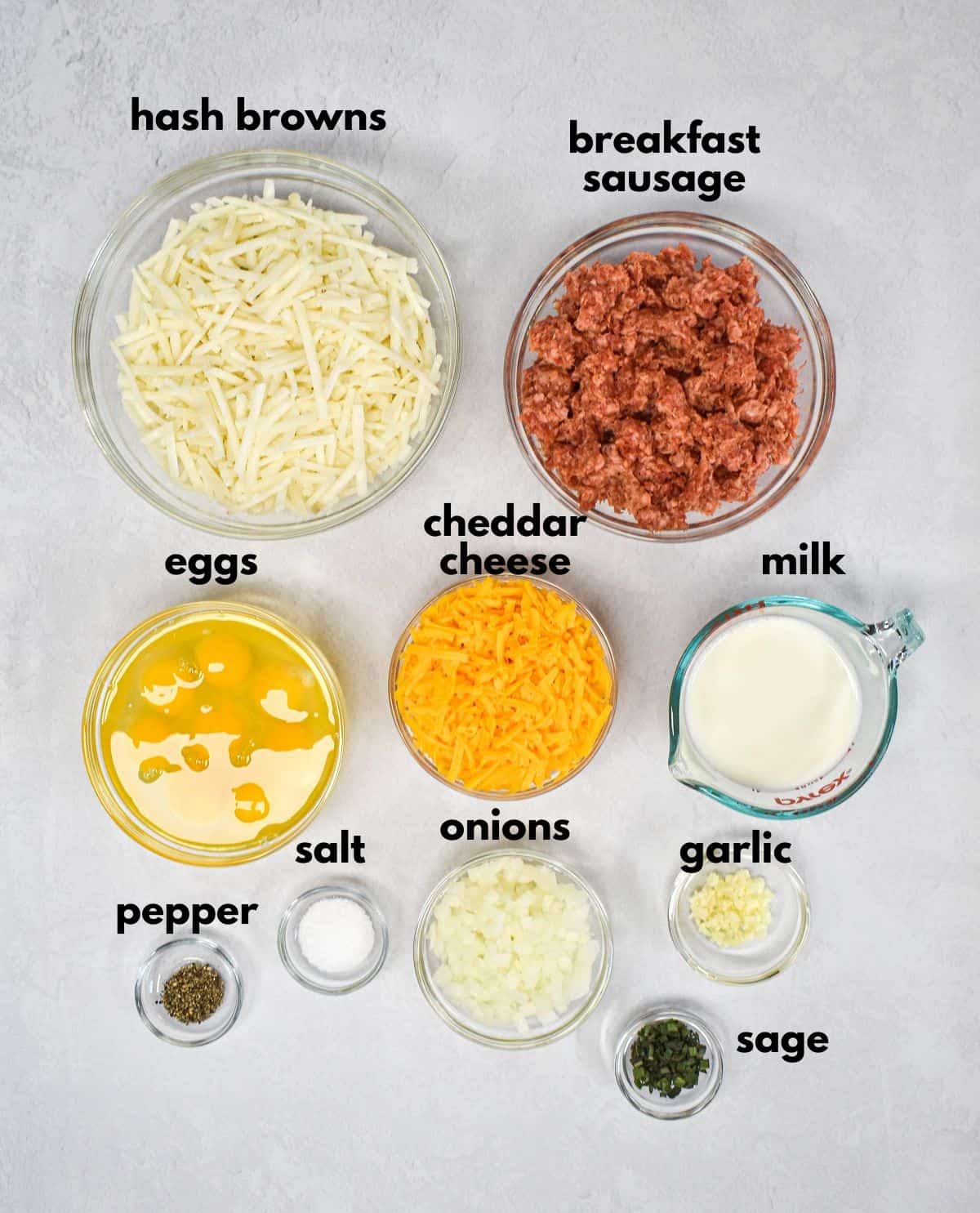 The ingredients for the breakfast casserole prepped and arranged in glass bowl on a white table with each labeled with small black letters.