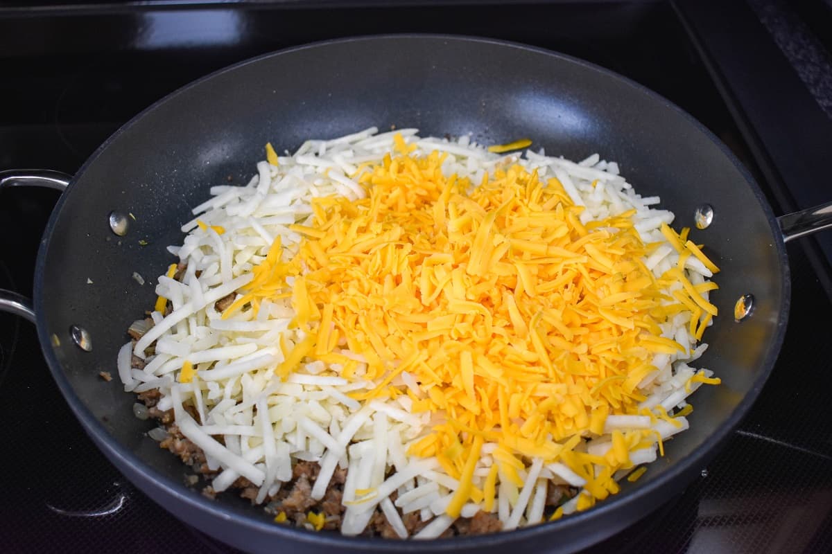 Hash browns and shredded cheese added on top of the browned sausage in a black skillet.