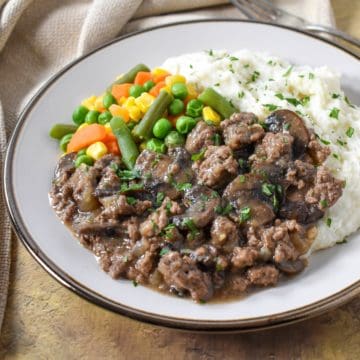 The ground beef and mushrooms served with mashed potatoes and mixed vegetables on a white plate.