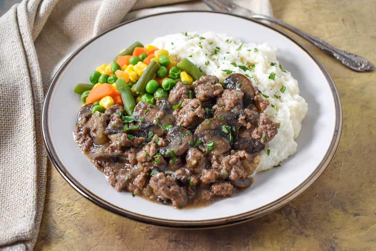 The ground beef and mushrooms served with mashed potatoes and mixed vegetables on a white plate.