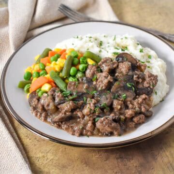 The ground beef and mushrooms served with mashed potatoes and mixed vegetables on a white plate.