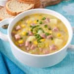 The corn and ham chowder served in a white bowl with bread slices in the background.