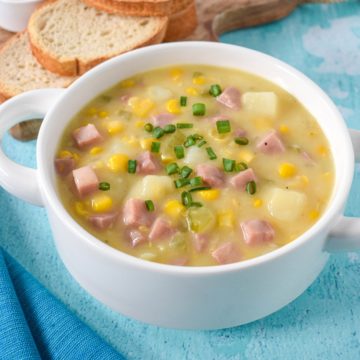 The corn and ham chowder served in a white bowl with bread slices in the background.