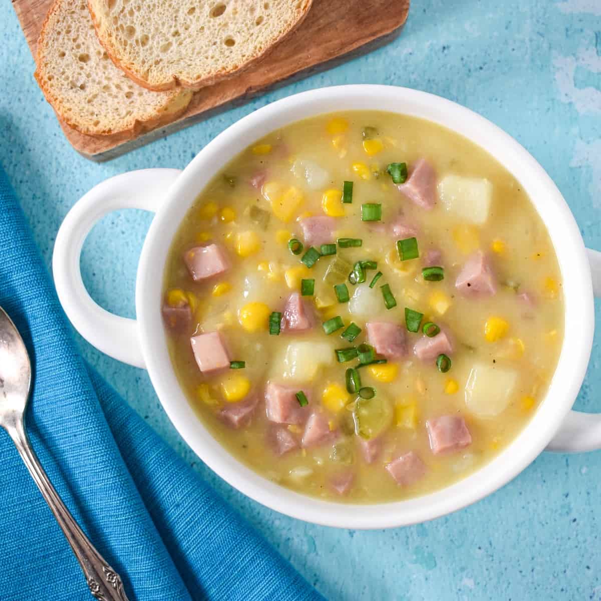 The corn and ham chowder served in a white bowl with bread slices in the background.