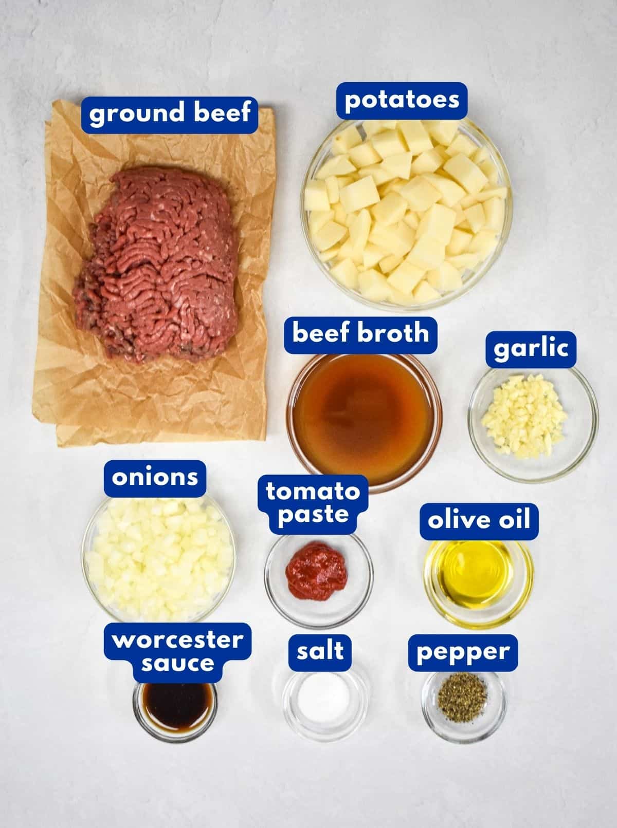 The ingredients for the ground beef and potatoes skillet prepped and arranged on a white table with each labeled in blue and white letters.