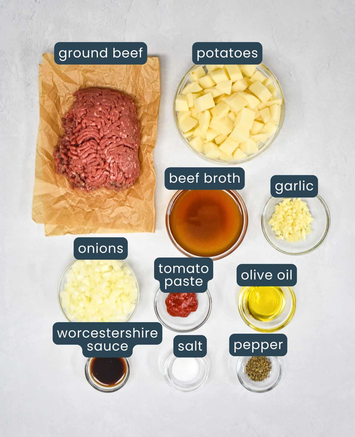 The ingredients for the ground beef and potatoes skillet prepped and arranged on a white table with each labeled in blue and white letters.