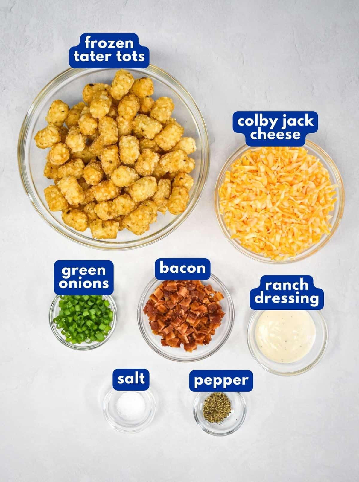The ingredients for the loaded tater tots prepped and arranged on a white table with each labeled in blue and white letters.