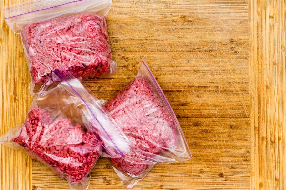Three zip top baggies with ground beef in them on a wood cutting board.