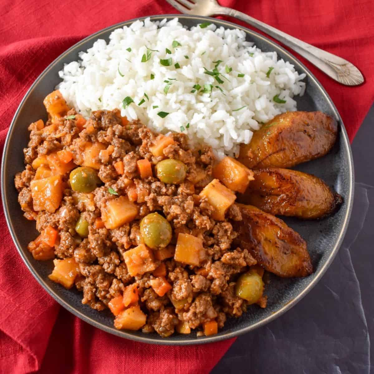 The picadillo served with white rice and fried sweet plantains on a gray plate.