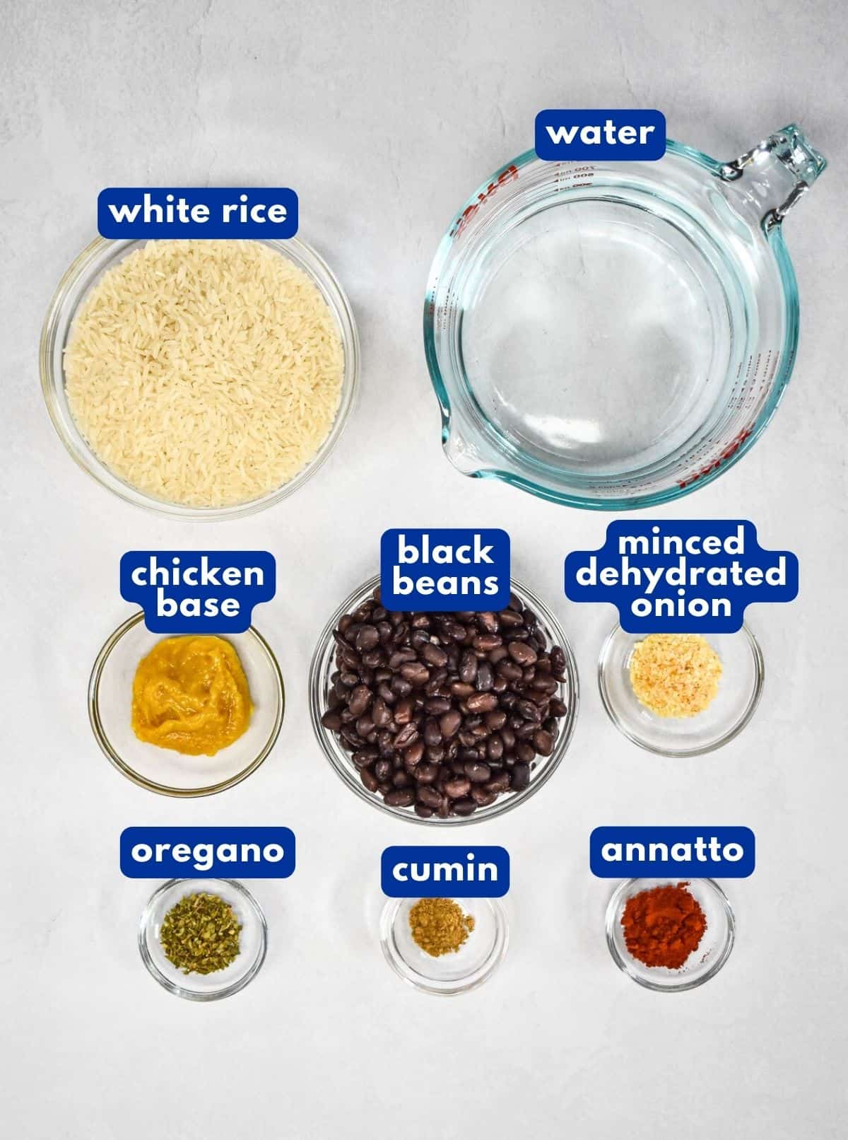 The ingredients for the rice dish prepped and arranged in glass bowls on a white table with each labeled with blue and white letters.