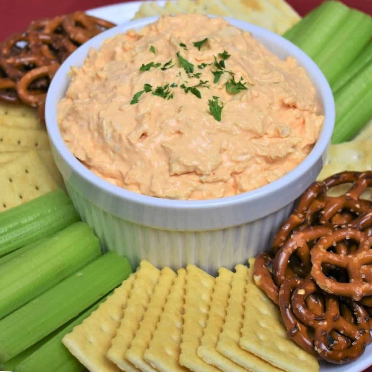 Cold Buffalo Chicken Dip served in a white bowl with assorted crackers, celery sticks and pretzels arranged on the plate.