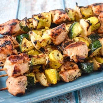 Skewers with grilled chicken, zucchini, yellow squash, and onions stacked on a blue plate.