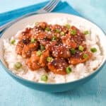Sesame chicken with chicken thighs served over a bed of white rice in a light blue and white bowl.