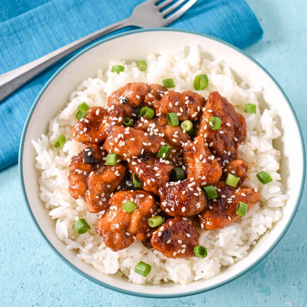 Sesame chicken with chicken thighs served over a bed of white rice in a light blue and white bowl.