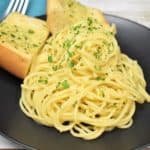 Spaghetti Aglio e Olio served on a black plate with two pieces of garlic toast in the background.