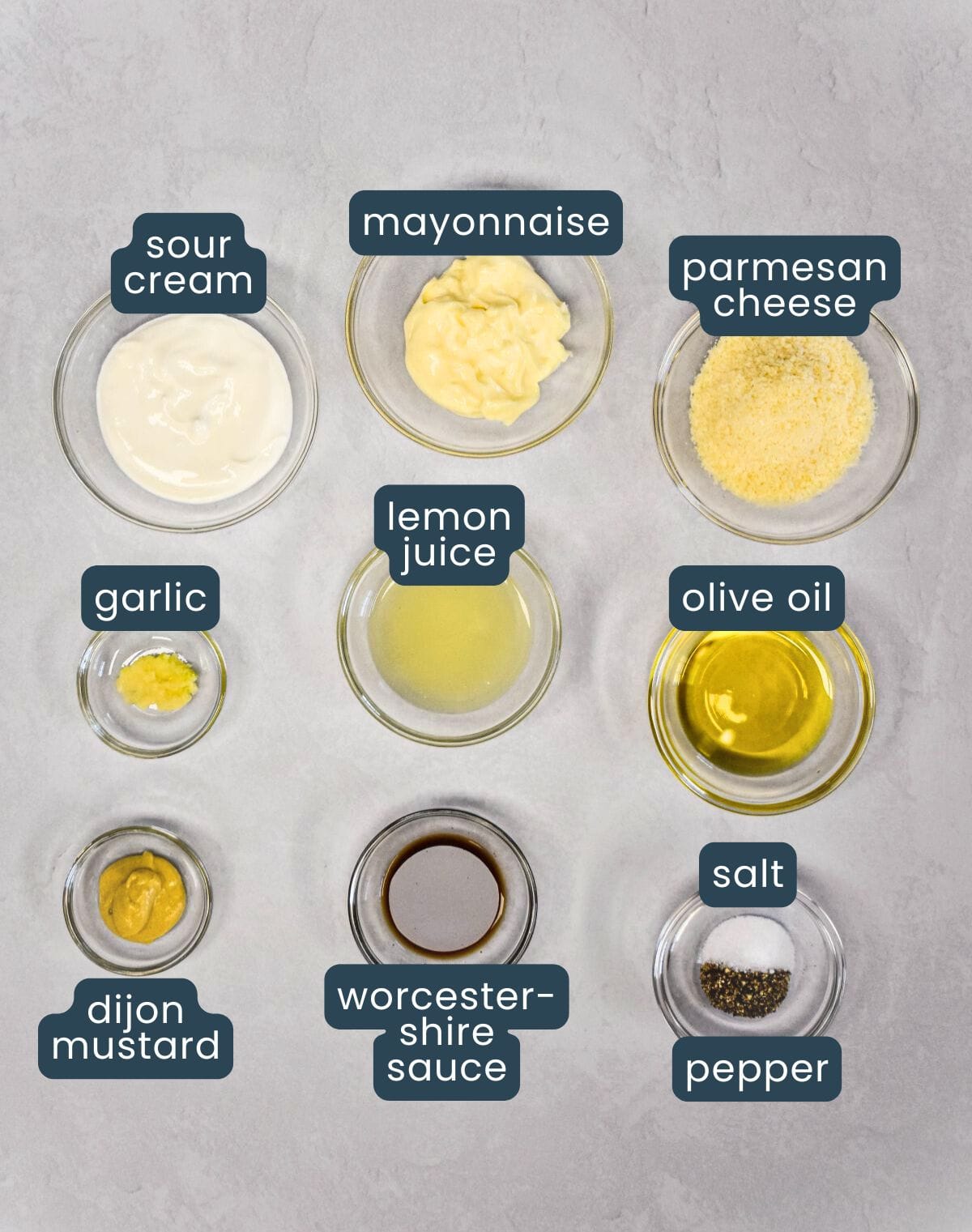Overhead shot of labeled ingredients in glass bowls, including mayonnaise, sour cream, lemon juice, Parmesan, and other components used to make the dressing.