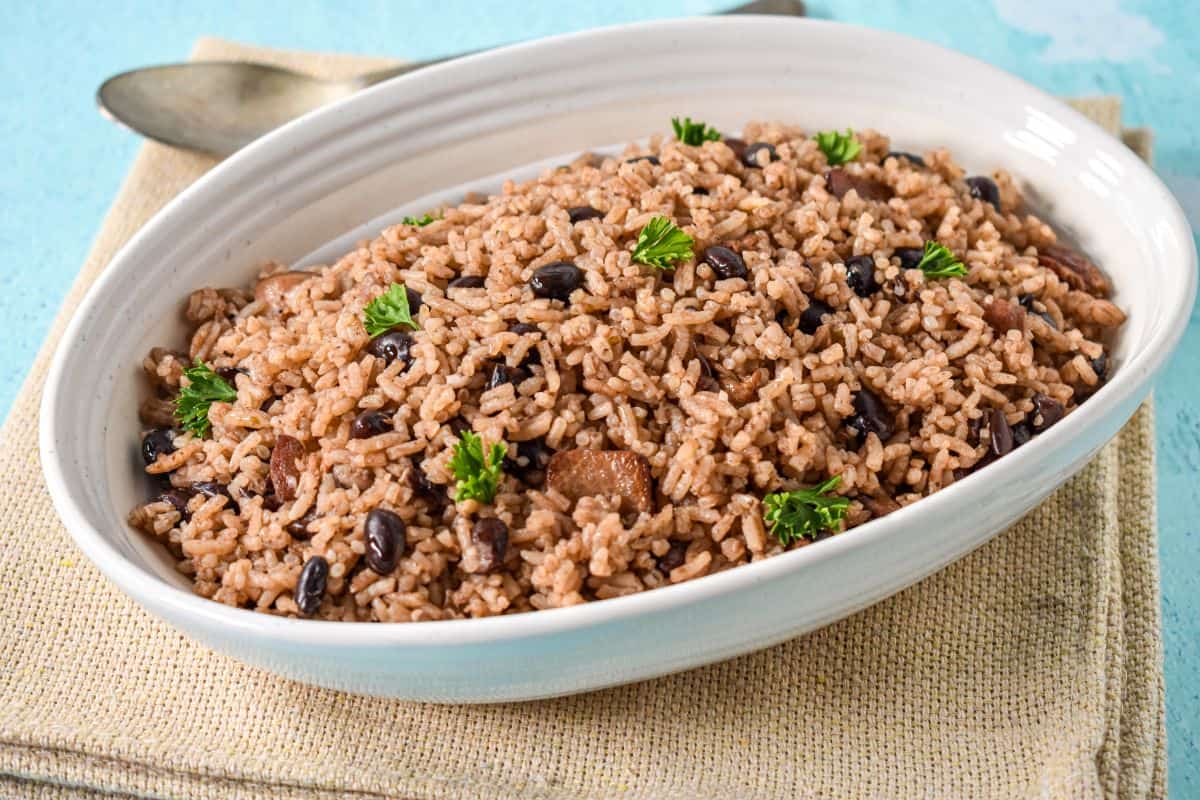 A white bowl filled with moros, showing black beans, rice, and bits of bacon. Garnished with parsley and set on a beige cloth napkin with a serving spoon in the background.
