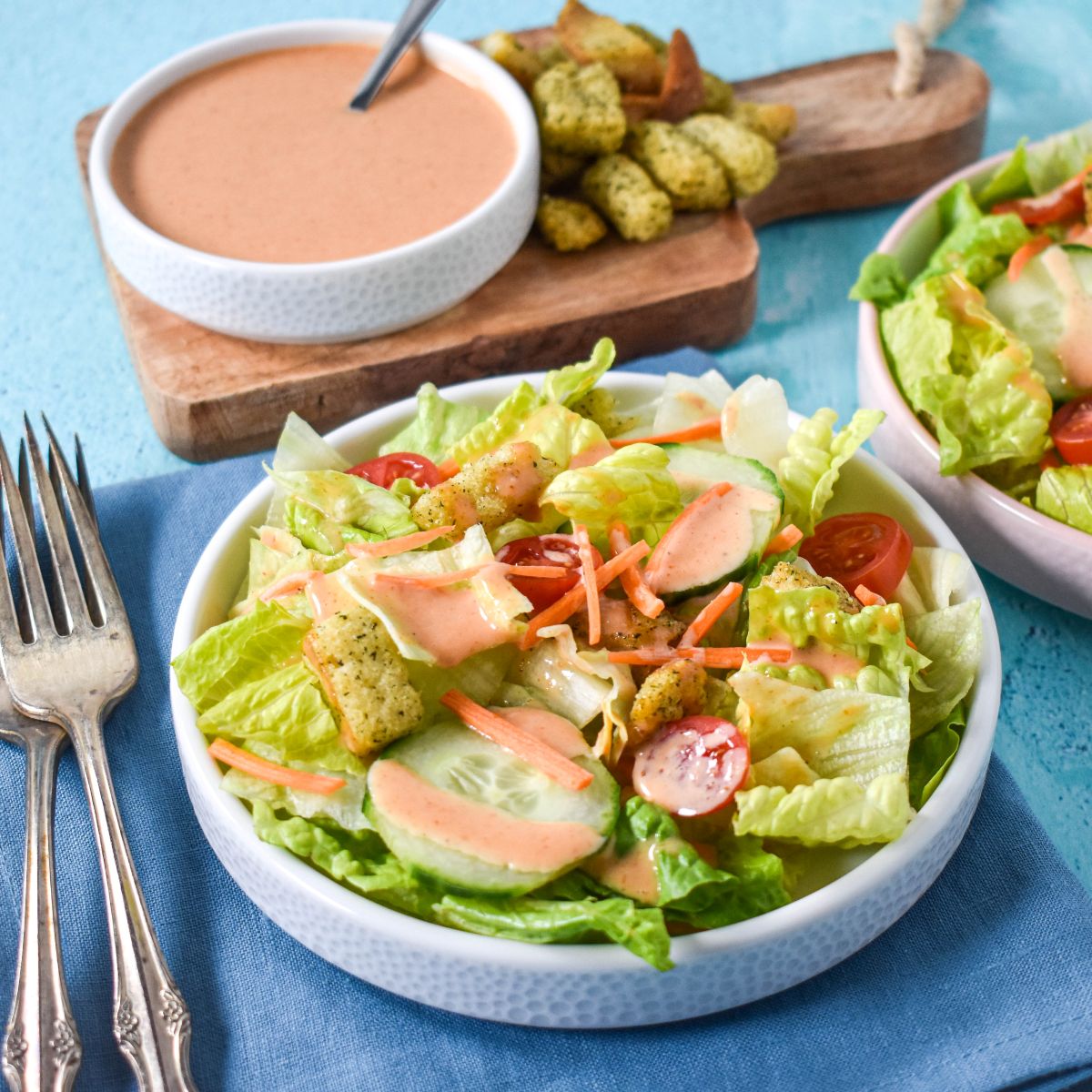 Side view of a salad topped with dressing and served with a bowl of dressing and croutons in the background.