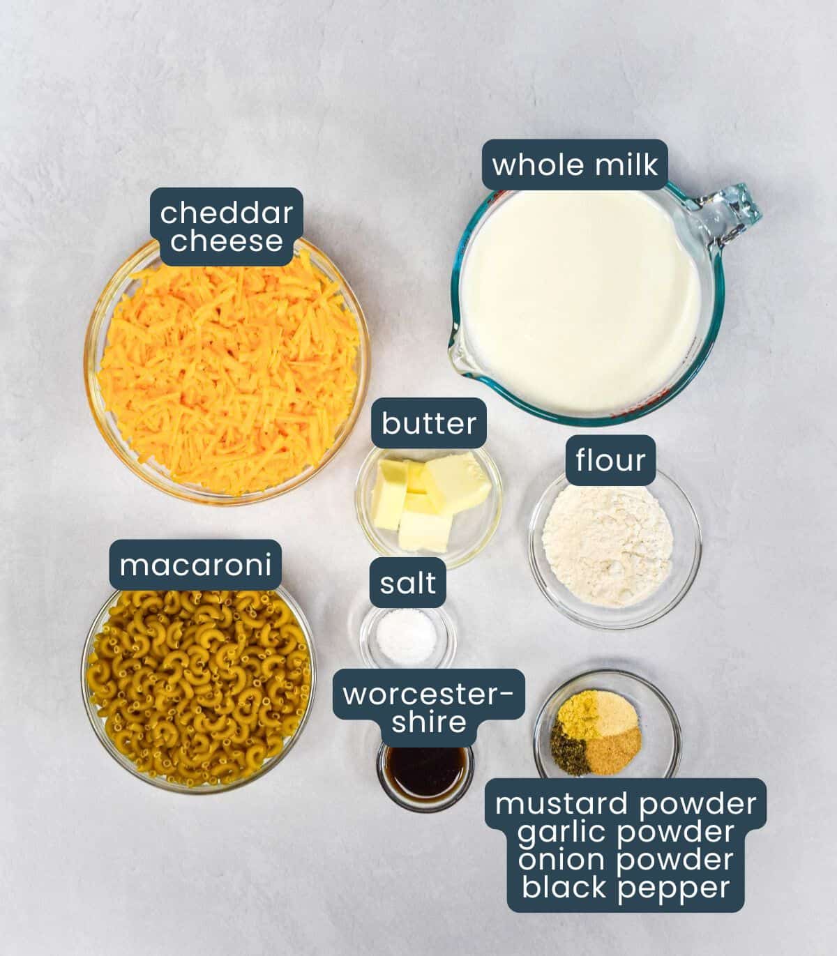 Overhead view of labeled ingredients in bowls, including shredded cheddar, macaroni, milk, butter, flour, salt, Worcestershire sauce, and spices.