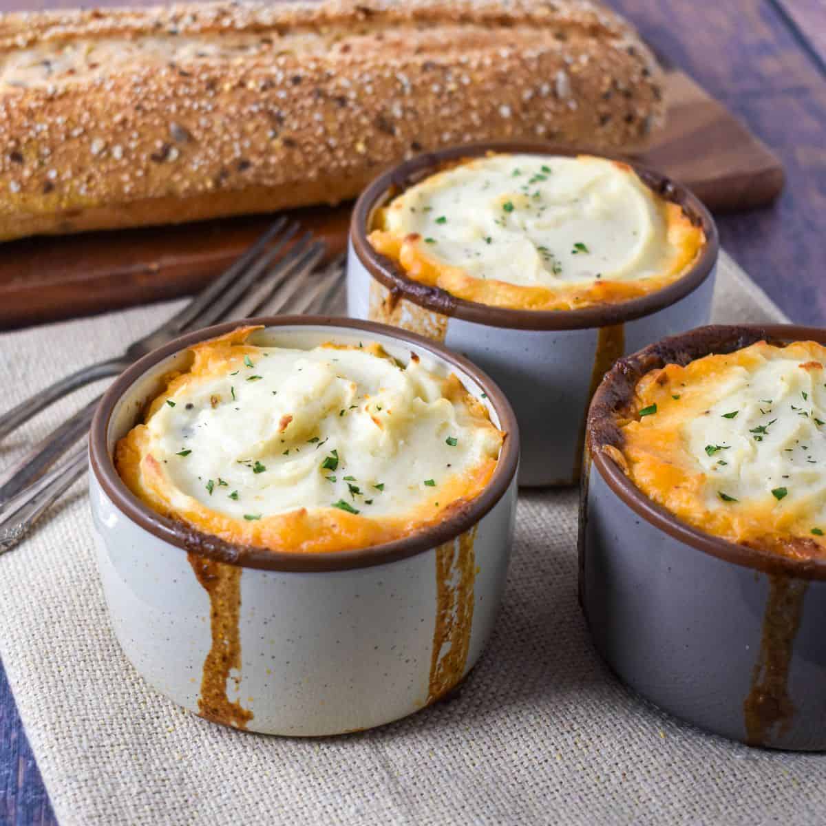 Three mini shepherd’s pies in ramekins on a table with forks and a loaf of multigrain bread in the background.