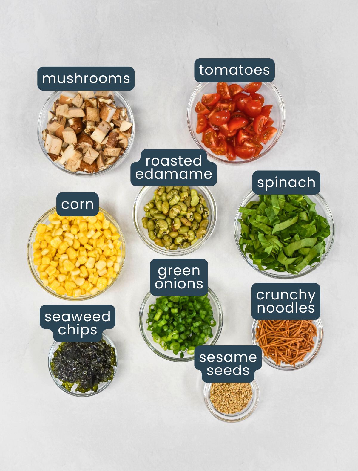 Labeled toppings for sesame ramen noodle salad, including tomatoes, mushrooms, corn, spinach, and seaweed chips.