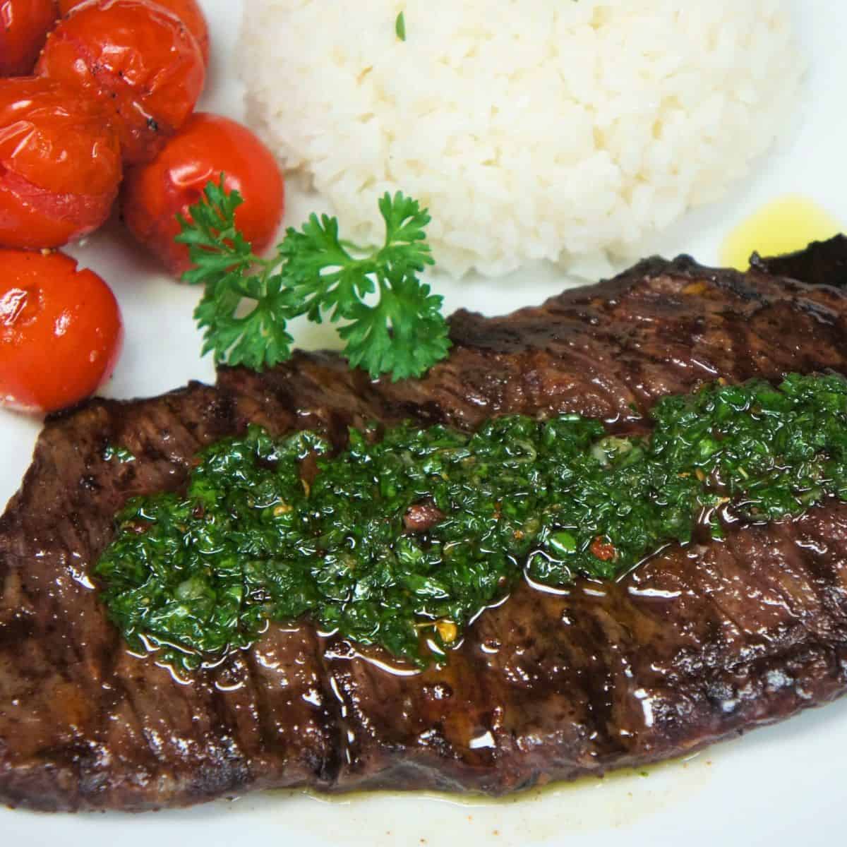 Cooked churrasco steak topped with chimichurri sauce and served on a white plate with rice and cherry tomatoes.