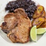 A pan fried pork chop served with white rice, black beans, fried sweet plantains and lime wedges on a white plate.