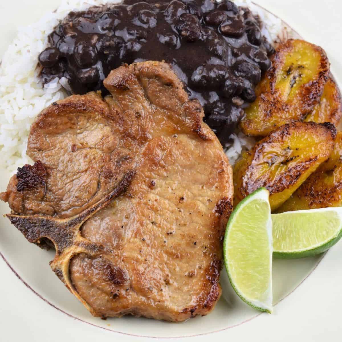 A pan fried pork chop served with white rice, black beans, fried sweet plantains and lime wedges on a white plate.