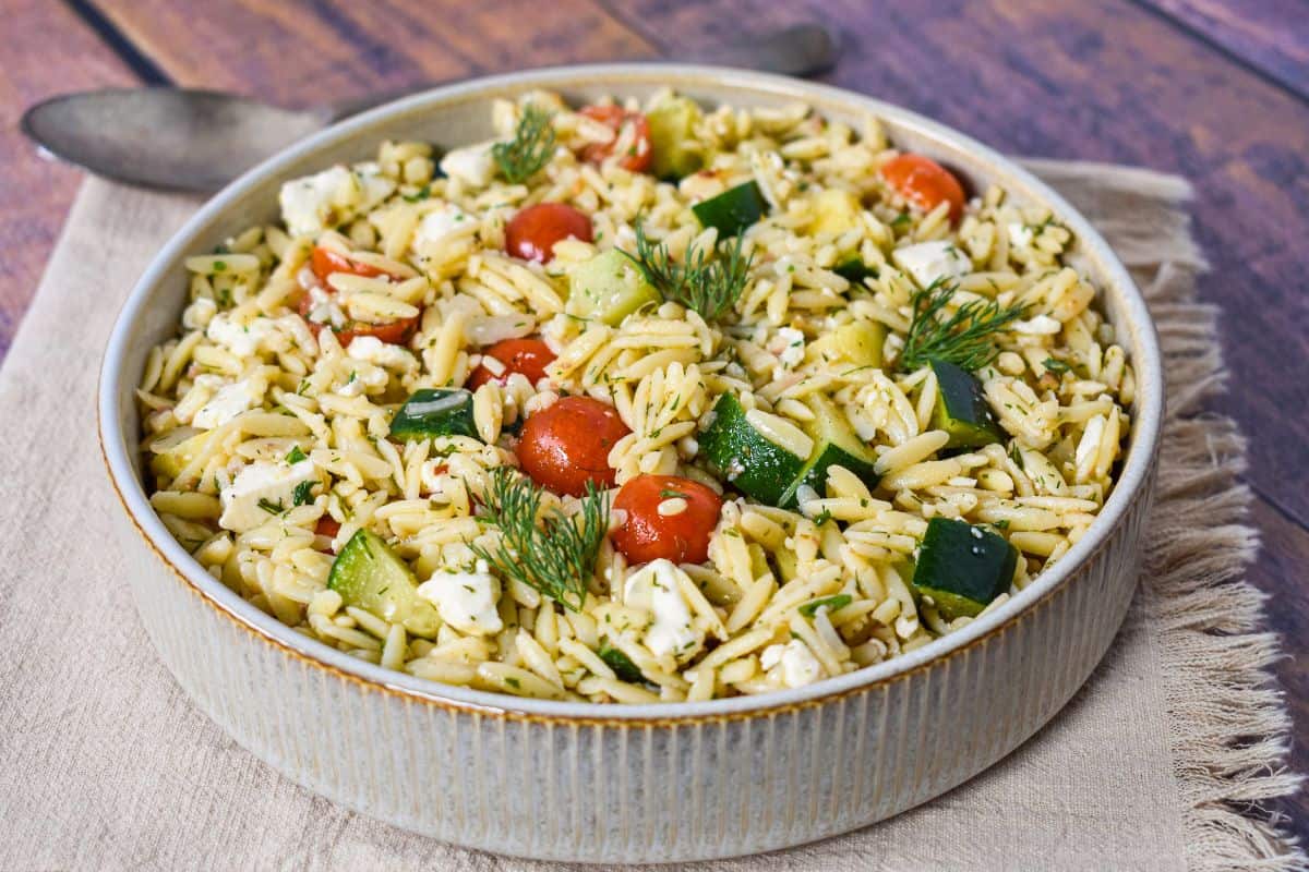 Angled view of herb orzo salad showing zucchini, tomatoes, feta, and fresh herbs in a ceramic bowl.