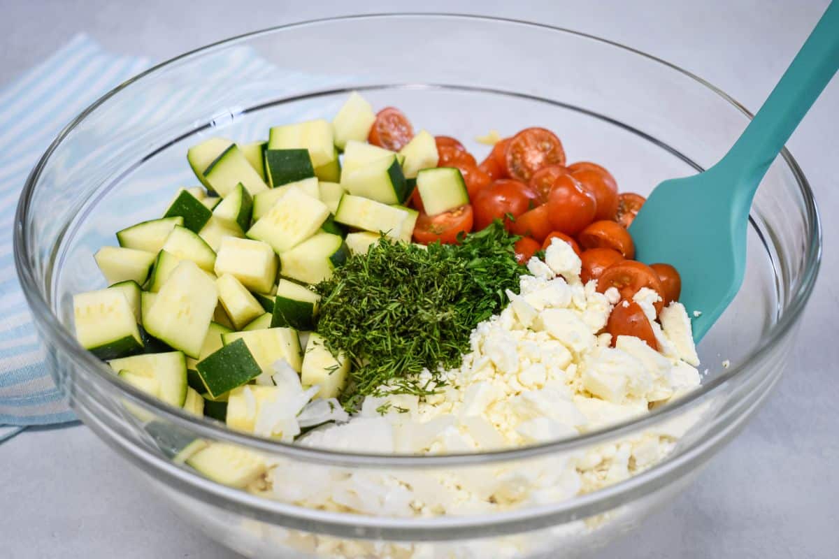 Ingredients for herb orzo salad in a mixing bowl before combining, including zucchini, tomatoes, feta, herbs, and onion.