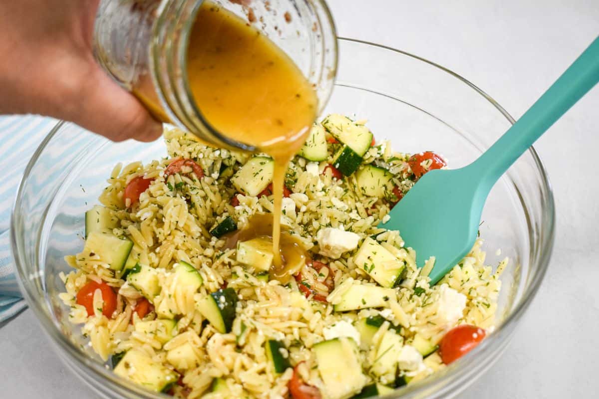 Italian dressing being poured over a bowl of herb orzo salad during preparation.