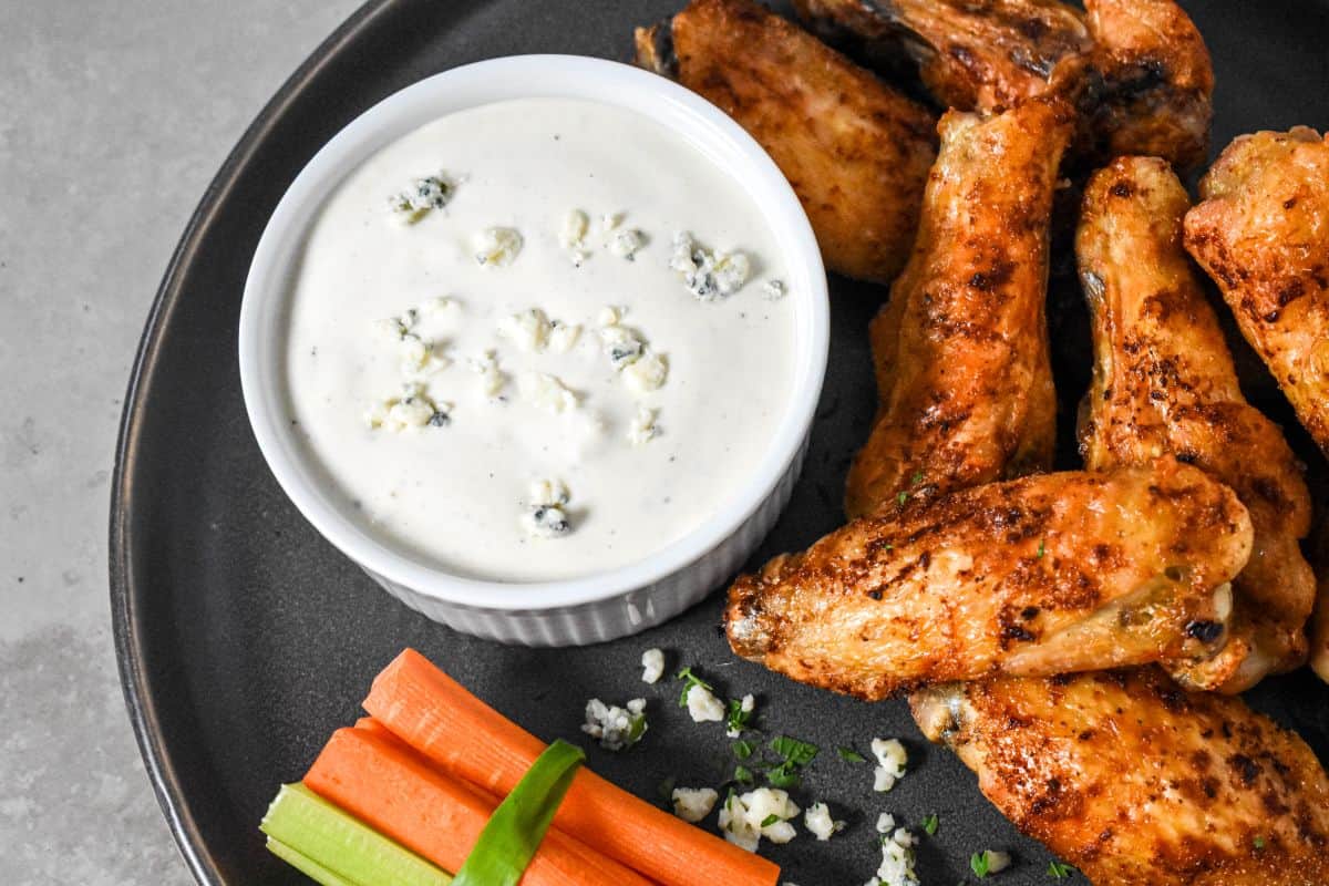 Close-up of chicken wings on a dark plate served with dressing on the side and scattered blue cheese crumbles.