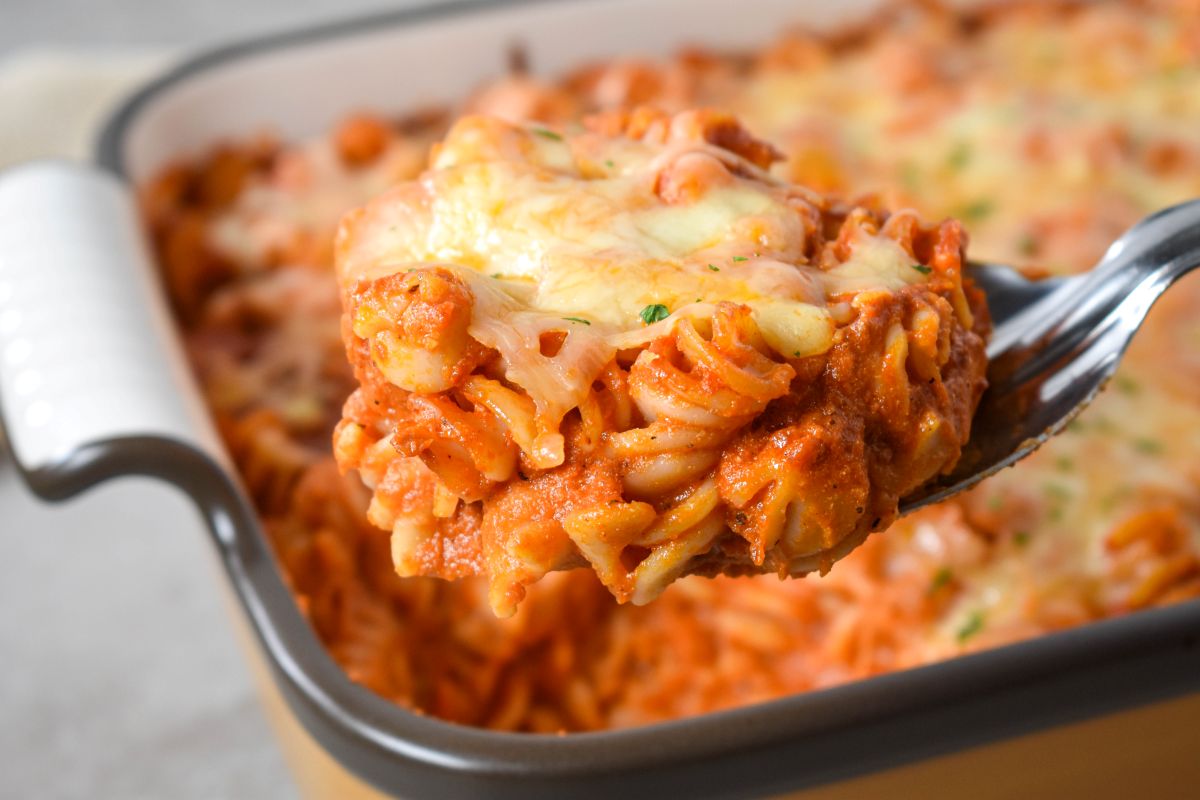 A close-up of a cheesy scoop of pasta lifted from the casserole dish, showing creamy sauce and melted cheese.