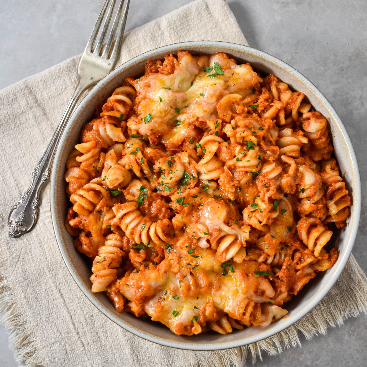 A bowl of vodka sauce pasta bake topped with melted cheese and parsley, served with a fork on a light napkin.