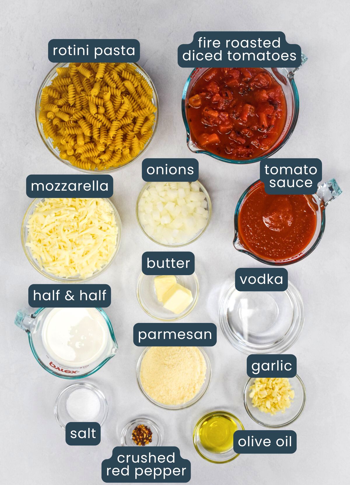 A labeled overhead image of ingredients in small bowls, including rotini pasta, fire roasted diced tomatoes, tomato sauce, onions, garlic, vodka, olive oil, butter, half and half, parmesan, mozzarella, salt, and crushed red pepper.