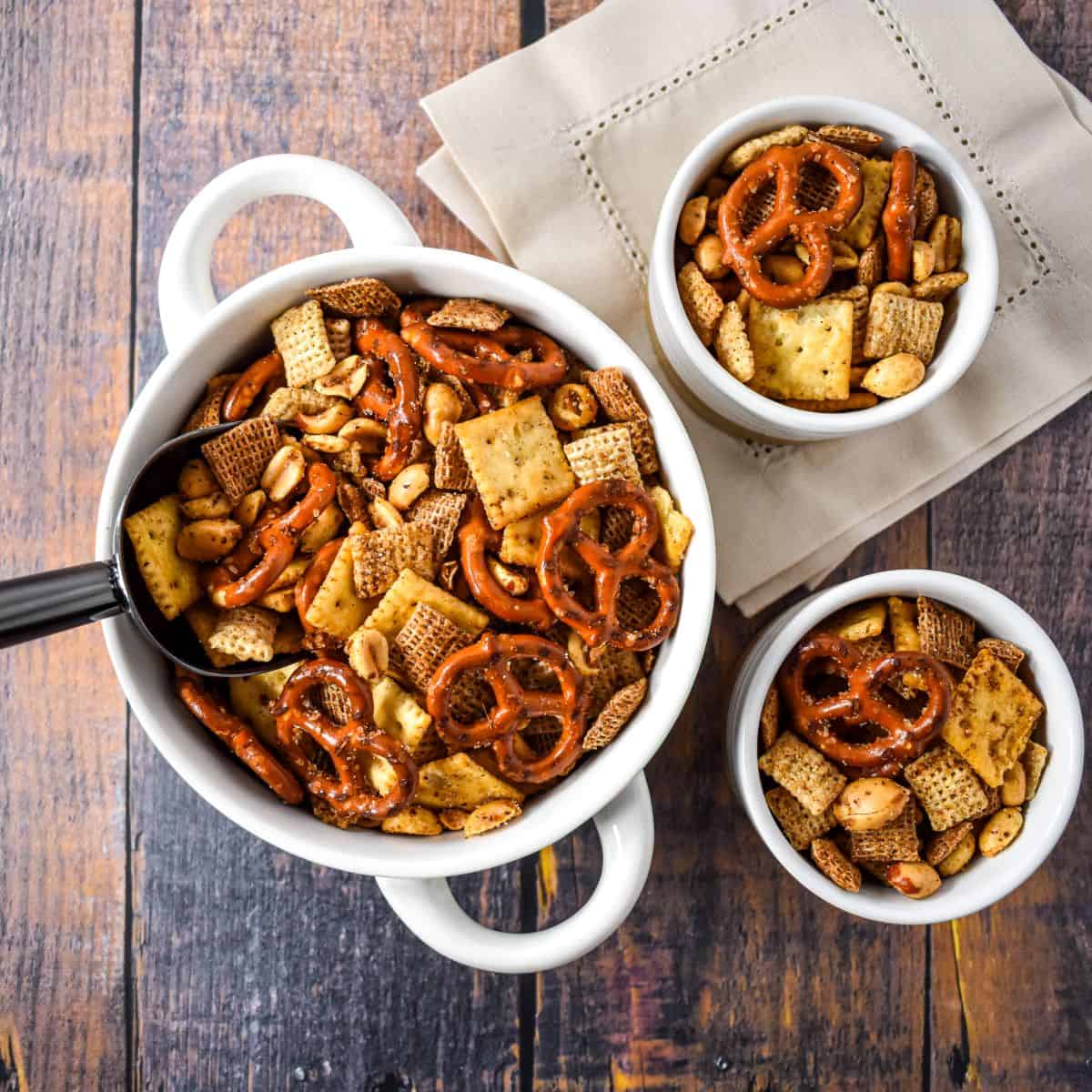 Cajun party mix in a large white serving bowl with smaller bowls on a wooden table.