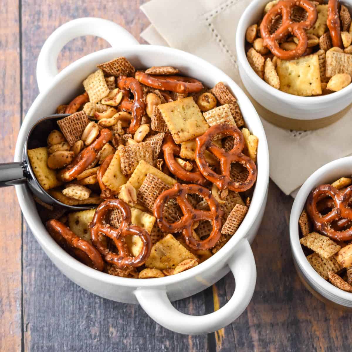 Close-up of Cajun party mix in a white bowl with a black scoop, next to smaller bowls filled with the snack.
