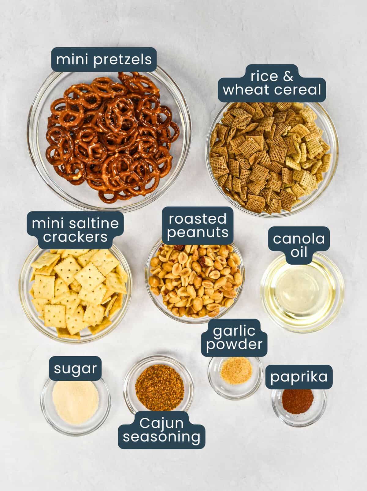 Glass bowls filled with pretzels, rice cereal, wheat cereal, mini saltine crackers, peanuts, and spices.