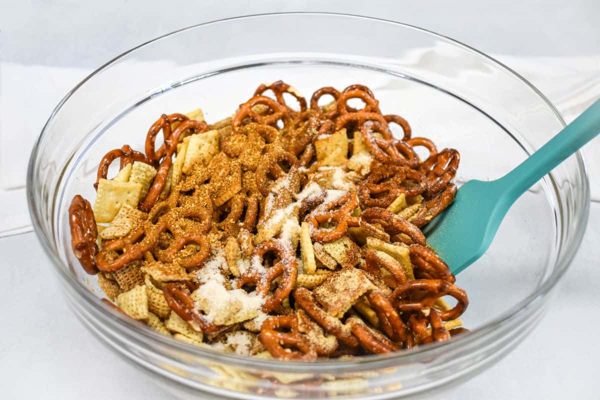 Seasoning blend of spices and sugar sprinkled over pretzels, crackers, and cereal in a mixing bowl.